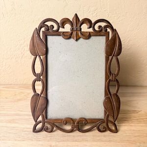 May include: Ornate brown metal picture frame with a fleur-de-lis at the top and leaf and chain design on the sides. The frame has a rectangular opening for a photo and sits on a wooden surface.