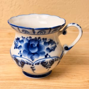 May include: A white ceramic cup featuring a scalloped rim and a rounded body decorated with hand-painted blue floral patterns. The design includes a central rose motif surrounded by intricate leaf details and a matching blue handle with accents.