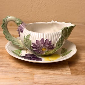 May include: A ceramic gravy boat and matching saucer featuring a white ribbed body with embossed purple flowers and green leaves. The handle is shaped like a leafy stem, and the saucer displays painted purple grapes and a yellow fruit design.