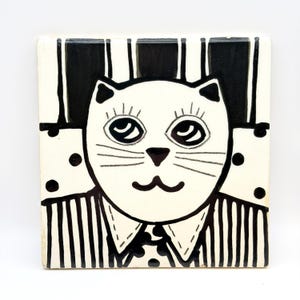 May include: Square ceramic tile featuring a whimsical cat design. The cat has a white face with black details, including large eyes, whiskers, and a small nose. The cat is wearing a striped shirt and a polka-dot tie. The background has a striped pattern and polka dots.