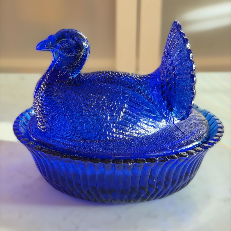 May include: Cobalt blue glass hen on nest. The hen is detailed with feathers and a curved neck, sitting atop a matching nest. The nest has a ribbed design and a decorative rim. A vintage-style decorative piece.