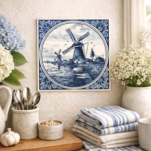 May include: Square blue and white ceramic tile featuring a windmill scene. The tile is framed with a floral border. A stack of blue and white striped towels and a vase of white flowers are in the foreground.