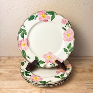 May include: A stack of white ceramic plates with a floral design. Each plate features pink flowers with yellow centers and green leaves around the rim. The plates are stacked on a wooden surface.