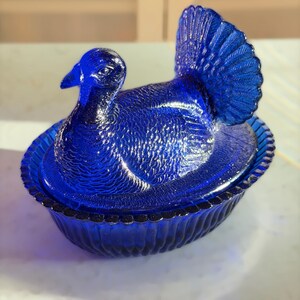 May include: A cobalt blue glass turkey-shaped trinket box. The lid features the turkey's head and fanned tail, while the base is a scalloped oval dish. The glass has a textured surface, adding depth to the design. A decorative piece.