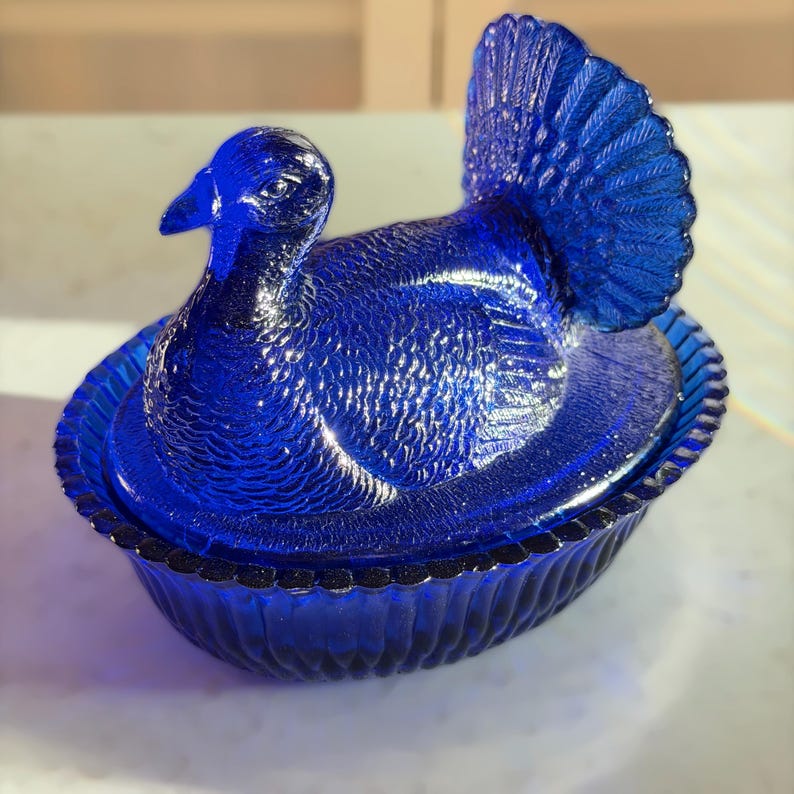 May include: A cobalt blue glass turkey-shaped trinket box. The lid features the turkey's head and fanned tail, while the base is a scalloped oval dish. The glass has a textured surface, adding depth to the design. A decorative piece.