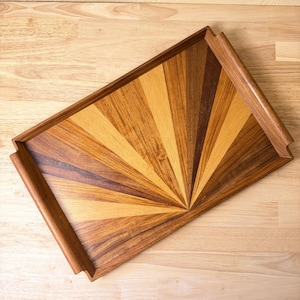 May include: A rectangular wooden serving tray featuring a sunburst pattern created from inlaid wood segments in varying shades of light tan, medium brown, and dark chocolate brown. The tray has raised side handles and a smooth finished surface.