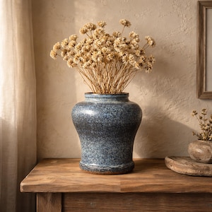 May include: A blue speckled ceramic vase with a wide base and a narrow neck, filled with dried beige flowers. The vase sits on a wooden table. The background features a neutral-toned wall and a framed artwork.