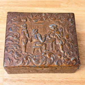 May include: A rectangular metal box with a dark, weathered bronze finish. The lid features a raised relief design depicting three figures in period clothing, with one seated at a table, surrounded by intricate, swirling floral scrollwork.
