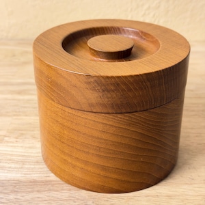 May include: A cylindrical wooden container with a smooth, polished finish and a visible natural grain pattern. The container features a matching circular lid with a small, centered knob handle on top, creating a uniform and cohesive appearance.