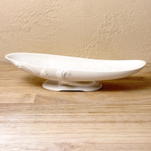 May include: A white, oval-shaped ceramic serving dish with a pedestal base. The dish has a gently curved design with decorative embossed floral details. The base is a solid, rounded shape. The dish is set on a wooden surface.