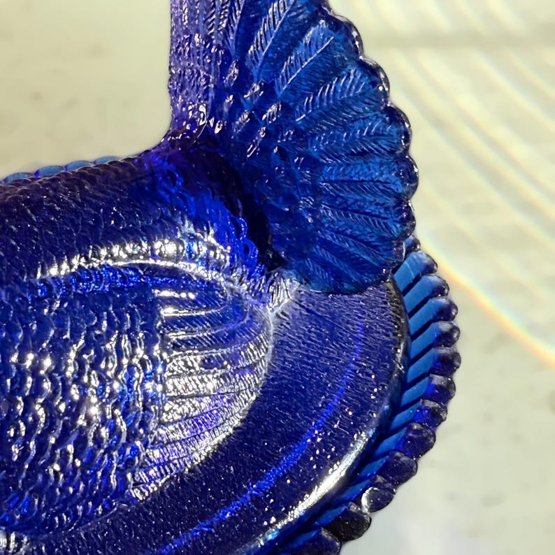 May include: Close-up of a cobalt blue glass decorative bird. The bird has textured details, including feathers and a braided edge. The glass reflects light, creating highlights and shadows.