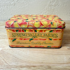 May include: Rectangular metal storage tin with a peach design on the lid. The sides feature the text "SPRING VALLEY FARMS Premium Quality Peaches" in green and brown. The tin is on a wooden surface.