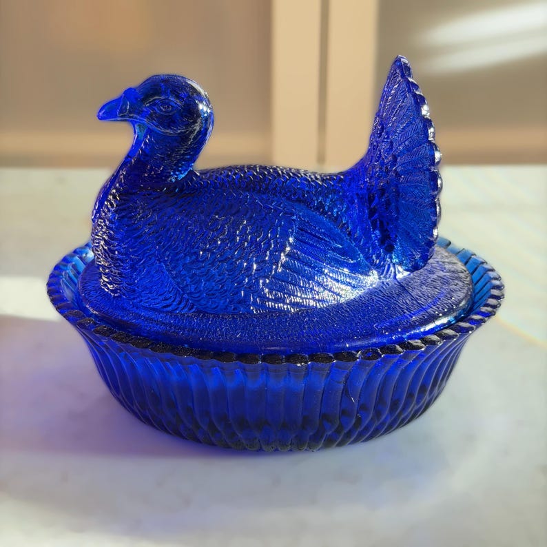 May include: A cobalt blue glass hen on a nest, a decorative dish. The hen is detailed with textured feathers and a curved neck. The nest is a matching blue with a ribbed design and a beaded rim. A vintage-style piece.