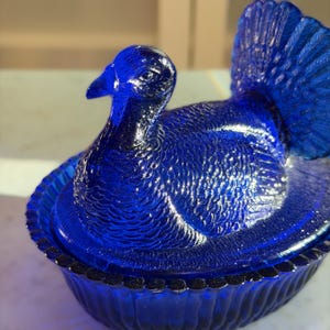 May include: A cobalt blue glass turkey figurine with detailed feather textures. The turkey is perched on a round, scalloped base. The bird's head is upright, and its tail feathers are fanned out. The glass has a glossy finish.