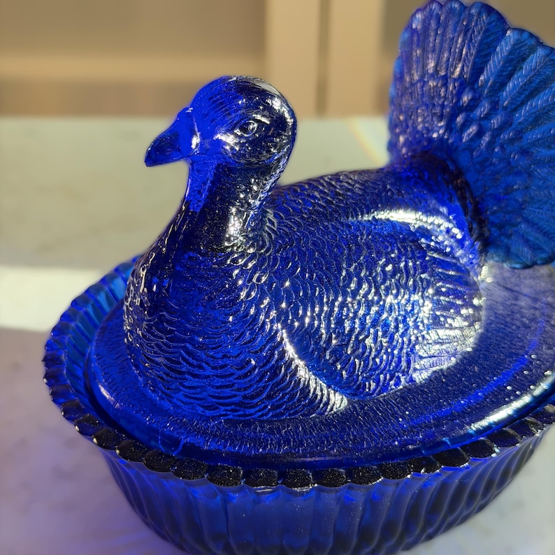 May include: A cobalt blue glass turkey figurine with detailed feather textures. The turkey is perched on a round, scalloped base. The bird's head is upright, and its tail feathers are fanned out. The glass has a glossy finish.