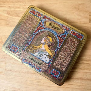 May include: A rectangular metal Whitman's Salmagundi tin featuring a colorful mosaic-style border in red, blue, and gold. The center displays an illustration of a person with long blonde hair, with the text Whitman's and Salmagundi printed.