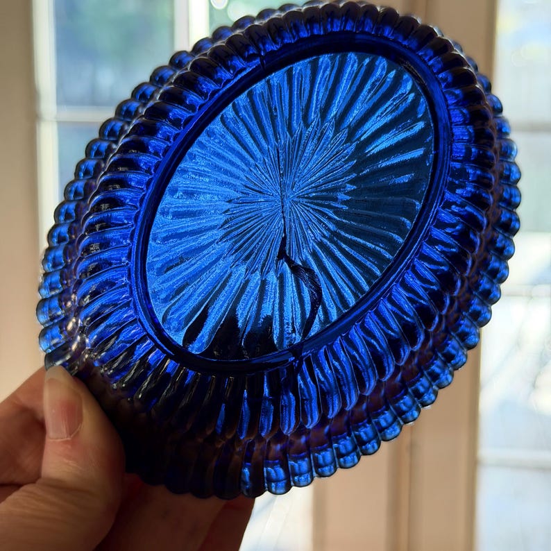May include: A vibrant cobalt blue glass dish with an oval shape. The dish features a textured, ribbed border and a central design of radiating lines. The glass has a glossy finish, reflecting light. The dish is held by a hand.