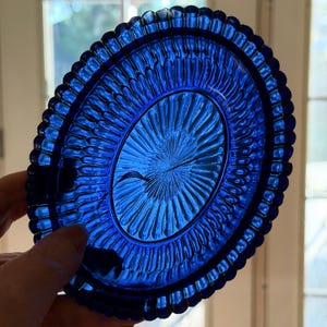 May include: A vibrant blue, oval-shaped glass dish. The dish features a detailed, textured pattern with a central design radiating outwards. The rim has a scalloped edge. The dish is held up by a hand.