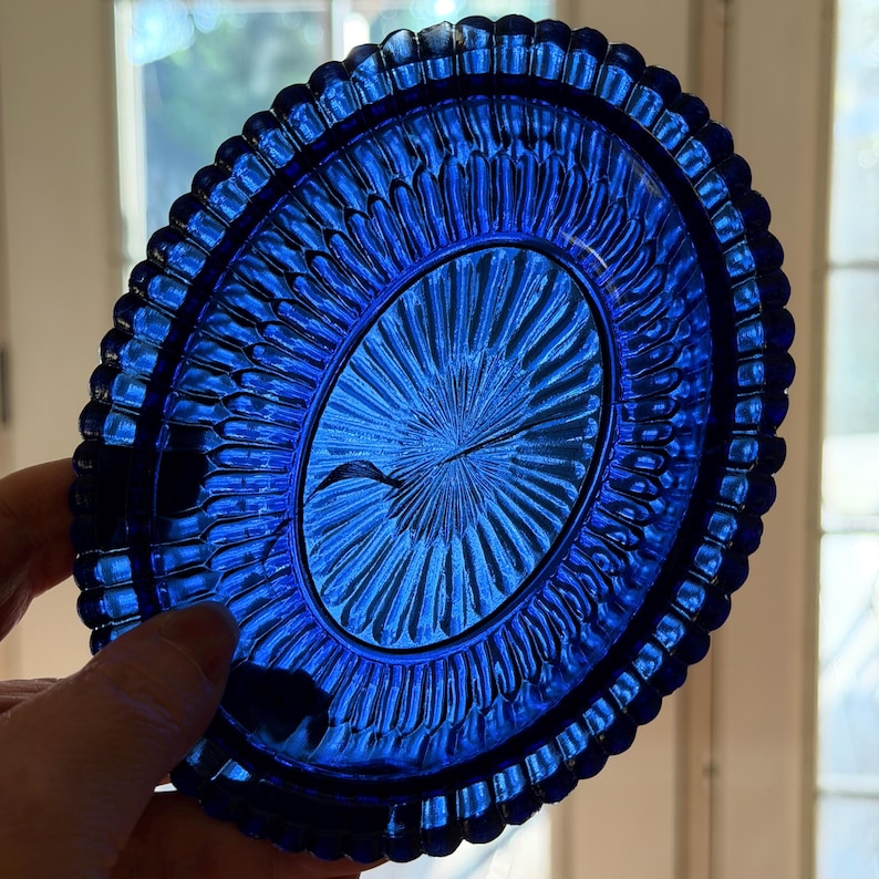 May include: A vibrant blue, oval-shaped glass dish. The dish features a detailed, textured pattern with a central design radiating outwards. The rim has a scalloped edge. The dish is held up by a hand.