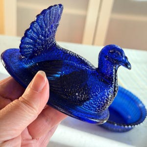 May include: A cobalt blue glass dish in the shape of a bird, with a detailed tail and head. The dish is open, revealing a separate, matching base. The bird's form is highly detailed, with textured feathers and a curved beak. The dish is held in a hand.