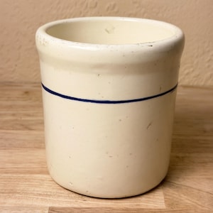 May include: A cylindrical off-white ceramic crock featuring a single thin horizontal navy blue stripe encircling the upper portion of the exterior. The vessel has a smooth, glazed finish with a thick, rounded rim and a flat base.