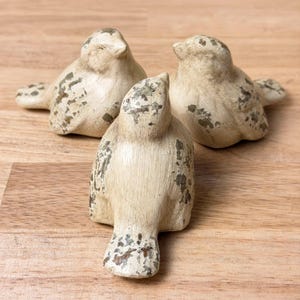 May include: Three small bird figurines crafted with a cream-colored base and a distressed, weathered finish featuring patches of dark gray and brown. Each bird is depicted in a seated position with wings tucked against the body and a short tail.