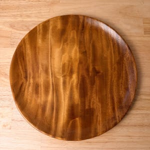 May include: A circular wooden plate featuring a smooth, polished surface with a prominent natural grain pattern in shades of medium brown and golden amber. The plate has a slightly raised rim and a consistent, warm-toned finish throughout.