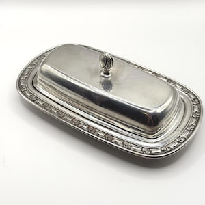 May include: A rectangular silver-toned butter dish with a removable lid. The base features a decorative border with a repeating floral pattern. The lid has a smooth, polished surface and a small, upright handle with an engraved design.