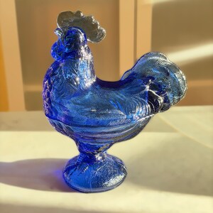 May include: A cobalt blue glass rooster figurine with a silver comb. The decorative piece has a detailed feather design and sits on a round base. The rooster is a decorative item.