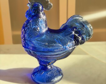 May include: A cobalt blue glass rooster figurine with a silver comb. The decorative piece has a detailed feather design and sits on a round base. The rooster is a decorative item.