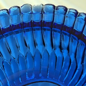 May include: Close-up of a vibrant blue glass bowl. The bowl features a repeating, textured pattern of vertical ridges, creating a play of light and shadow. The rim has a scalloped edge, adding to its decorative appeal. The bowl's color is a deep, saturated blue.