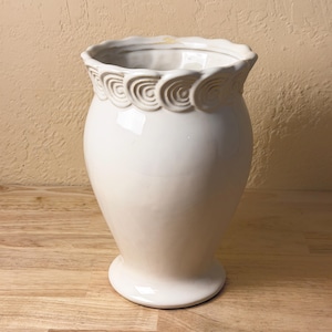 May include: A glossy off-white ceramic vase with a wide, scalloped rim featuring a repeating embossed spiral pattern. The vase has a bulbous body that tapers down to a circular pedestal base, presenting a smooth, reflective surface throughout.