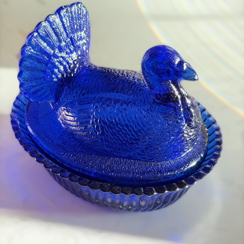 May include: A cobalt blue glass turkey-shaped dish with a lid. The turkey has detailed feather and body textures. The dish has a scalloped edge and is designed to hold food. A decorative piece for the kitchen or dining room.