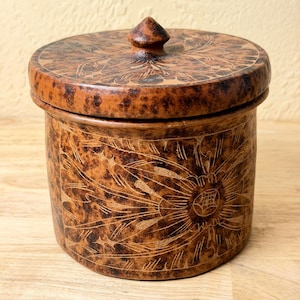 May include: A cylindrical brown lidded container with a mottled, dark-spotted finish. The surface features intricate, etched floral patterns across the body and the lid, which is topped with a small, pointed, central handle knob.