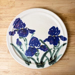 May include: A round, white ceramic plate with a raised edge. The plate is decorated with hand-painted blue iris flowers with purple accents and green stems and leaves. The plate is set on a light wood surface.
