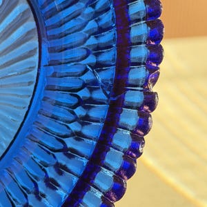 May include: Close-up of a vibrant blue glass plate with a textured, ridged design. The plate's edge features a scalloped pattern, creating a decorative and eye-catching aesthetic. The color is a deep, rich blue.