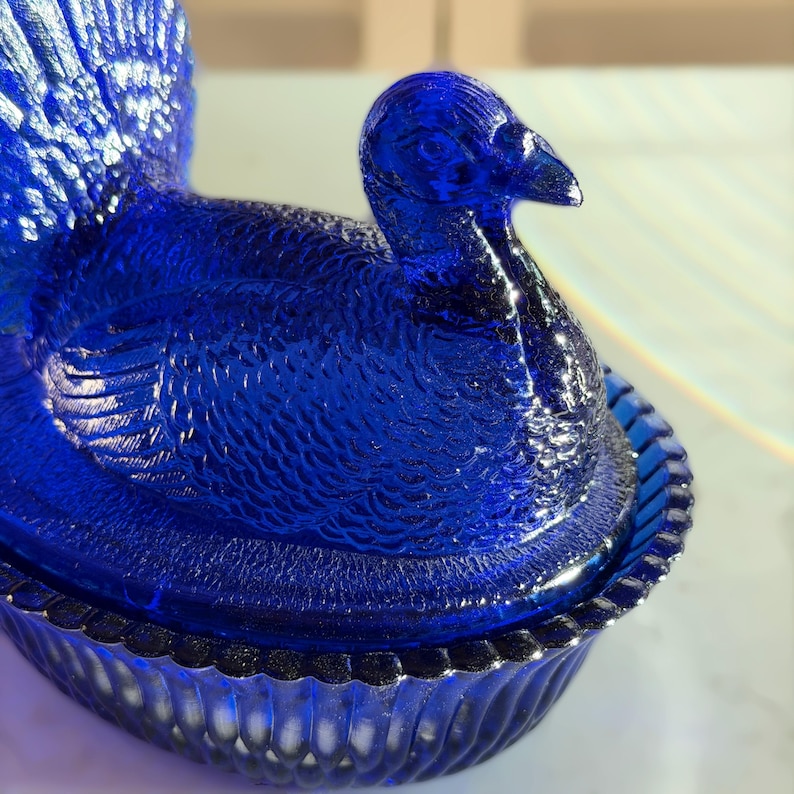 May include: A cobalt blue glass turkey-shaped dish with a textured surface. The turkey has a detailed feather design and a decorative rim. The dish is likely vintage and suitable for serving or display.
