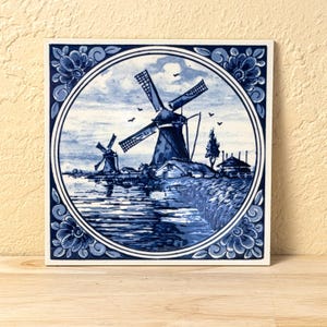 May include: Square ceramic tile featuring a blue and white windmill scene. The image depicts two windmills near water, with a circular border and floral accents. The tile is displayed on a wooden surface.