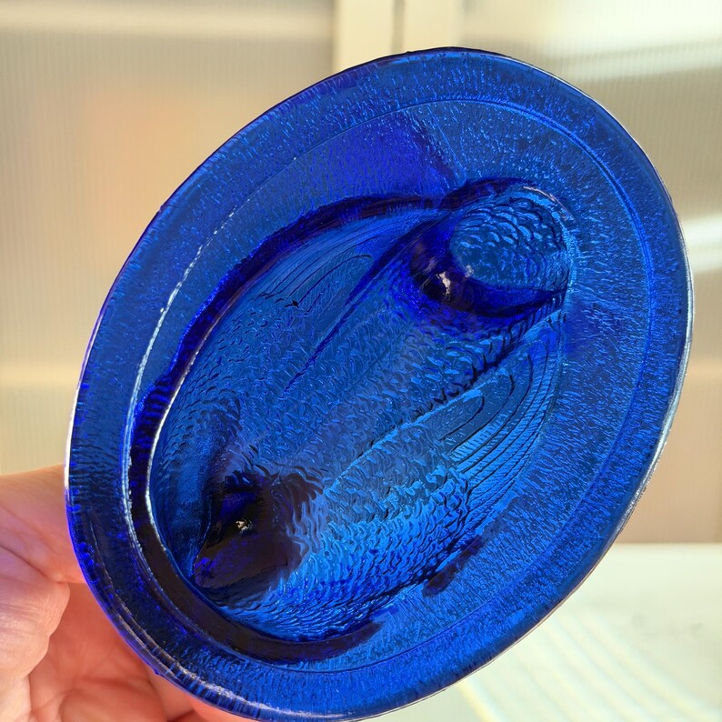 May include: A cobalt blue, oval-shaped glass dish with a textured surface. The dish features a raised relief design of a bird with outstretched wings. The bird's feathers and details are visible in the glass.