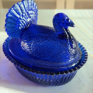 May include: A cobalt blue glass turkey-shaped dish with a lid. The turkey has detailed feather and body textures. The dish has a scalloped edge and is likely used for serving or decoration. The glass has a glossy finish.