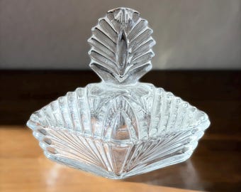 May include: A clear, vintage glass dish with a lid. The dish has a fan-like pattern and a decorative, leaf-shaped handle on the lid. The dish is likely used for serving or storing small items.