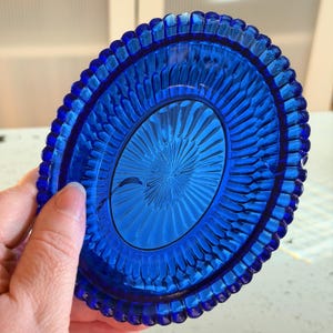 May include: A vibrant blue glass dish with a detailed, textured design. The dish has an oval center with a radiating pattern and a scalloped edge. The color is a deep, rich blue, and the dish is held by a hand.