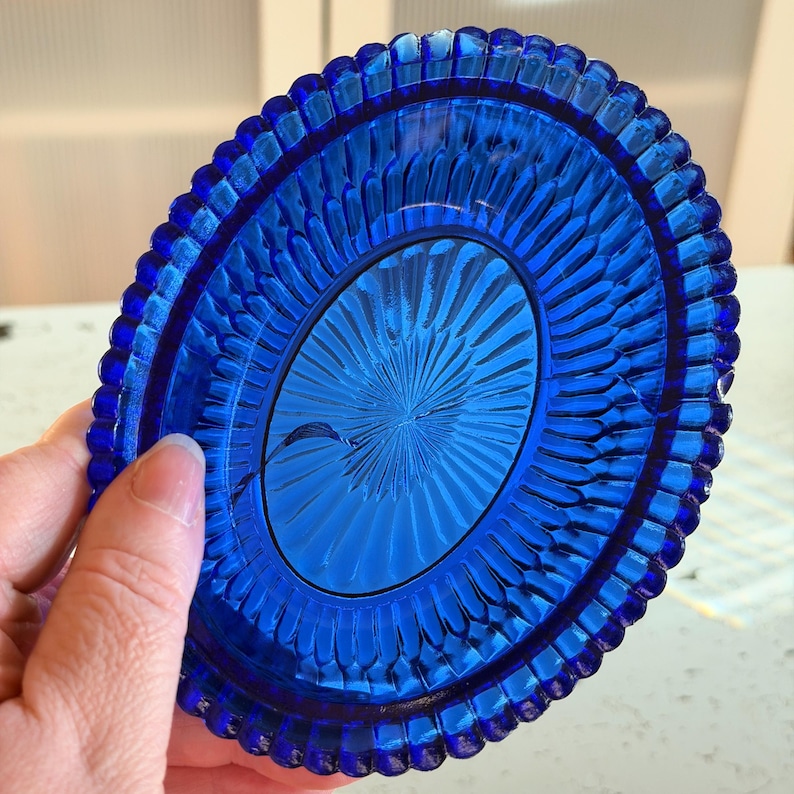 May include: A vibrant blue glass dish with a detailed, textured design. The dish has an oval center with a radiating pattern and a scalloped edge. The color is a deep, rich blue, and the dish is held by a hand.