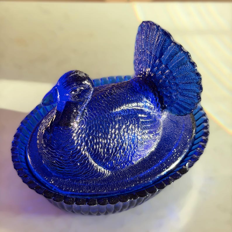 May include: A cobalt blue glass turkey-shaped dish with a textured surface. The dish features a detailed turkey design with a fanned tail and sits in a decorative oval base. The lid is also shaped like a turkey.