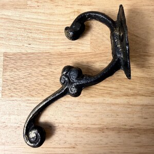 May include: A black cast iron wall hook featuring a double-pronged design with a curved upper hook and a lower hook ending in a decorative scroll. The mounting plate is rectangular with visible screw holes for attachment to a flat surface.