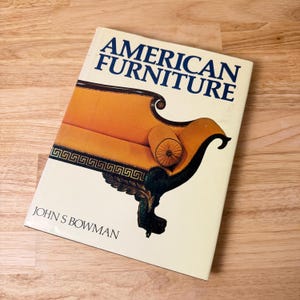 May include: A book titled "AMERICAN FURNITURE" by John S. Bowman. The cover features an illustration of an orange sofa with dark brown and black accents. The sofa has a decorative design and a matching bolster pillow.