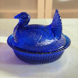 May include: A cobalt blue glass duck-shaped dish with a lid. The duck is detailed with textured feathers and sits in a ribbed bowl. The lid forms the duck's body and tail. A decorative piece for the home.