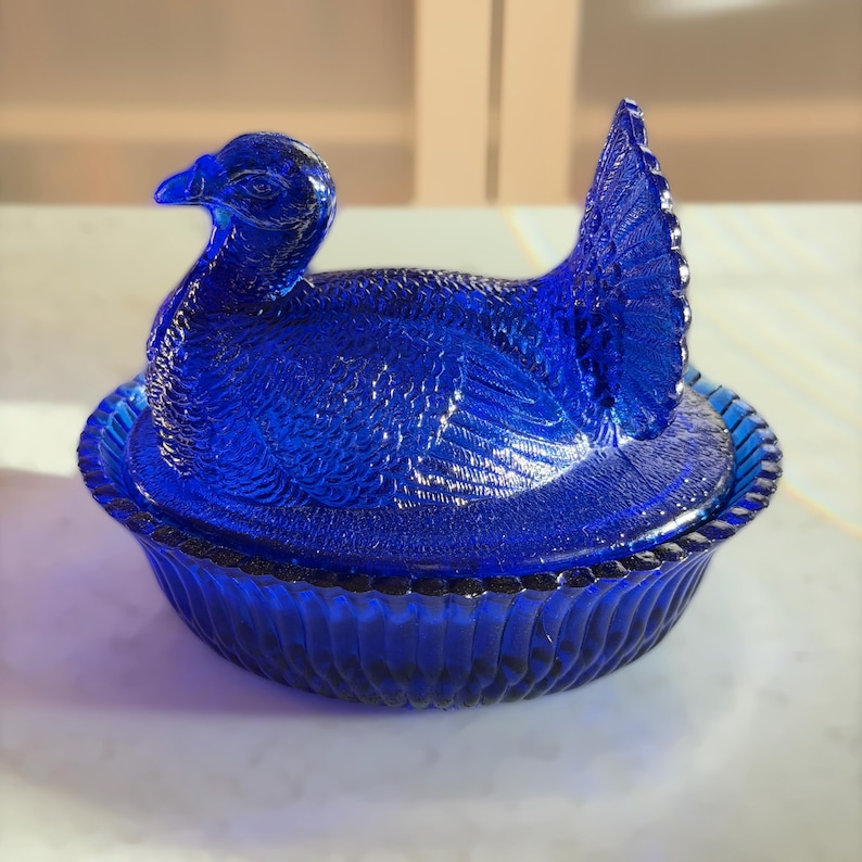 May include: A cobalt blue glass duck-shaped dish with a lid. The duck is detailed with textured feathers and sits in a ribbed bowl. The lid forms the duck's body and tail. A decorative piece for the home.