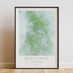 May include: A framed print of the Peak District National Park in England. The artwork features a green and white topographical map design. The print is in a black frame and leans against a white wall on a wooden floor.