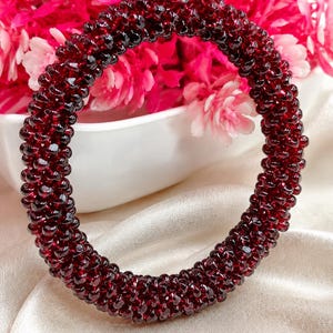 May include: A circular bangle bracelet constructed from numerous small, deep red, faceted garnet beads. The beads are densely packed together to form a continuous, textured, and rounded band that reflects light across its dark crimson surface.